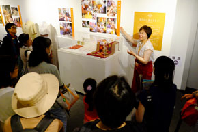 ショートカットの女性が、展示ケースの横で、取り囲む人々に向かって話している。