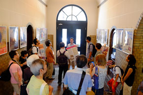 趣のある建物内部で、こちらに向かって立つ一人の男性と、男性を取り巻く十数人の人々。