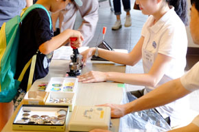 赤い柄の付いた道具に手を掛ける男児。向かい側に座る大人。手前には金属の部品が見える。