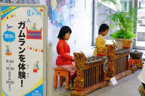 「ガムランを体験！」と書かれた看板の横で、鍵盤楽器を演奏する二人の少女。
