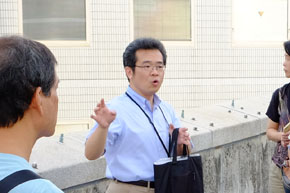 身振りを交えながら熱心に話す眼鏡を掛けた男性。両側に聴衆が二人写り混んでいる。