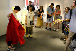 赤い布を広げて身にまとおうとする女性と、それを見守る人々。