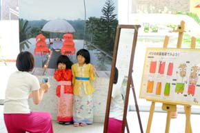 色鮮やかな衣装を着て風景写真の前でポーズをとる二人の幼女と幼女に向かってカメラを構える女性。