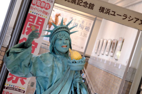 開館祭のバナーの前で歌を歌うかのようなポーズをとる、顔や服を水色に塗り冠を被った女性。