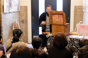 紙芝居を見せる眼鏡の男性と、見入る人々。