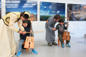 椅子に座って馬頭琴を持つ2人の男の子。モンゴル服の女性2人が、弓の持ち方や弾き方を教えている。