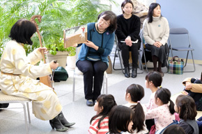 絵本を掲げて挿絵を見せる女性。隣りに馬頭琴を持った女性。絵本に見入る子どもたち。